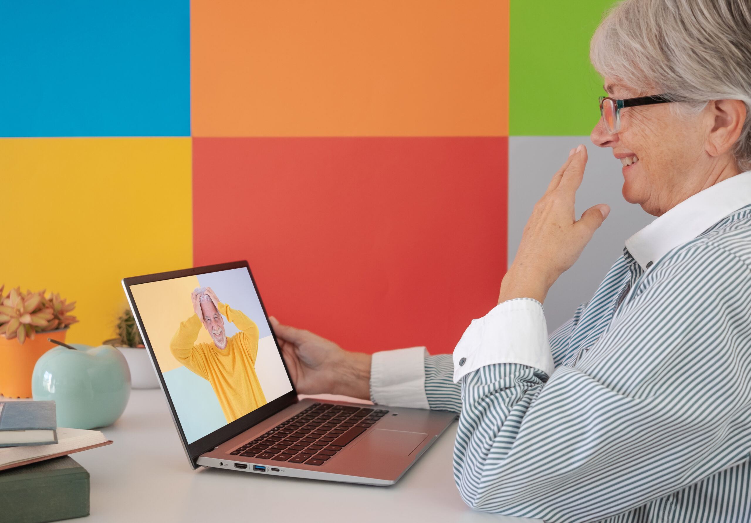 Elderly woman amused video chatting with a friend by laptop. Mature woman laughing while she talks to a distant man with his hands in his hair. Colored background