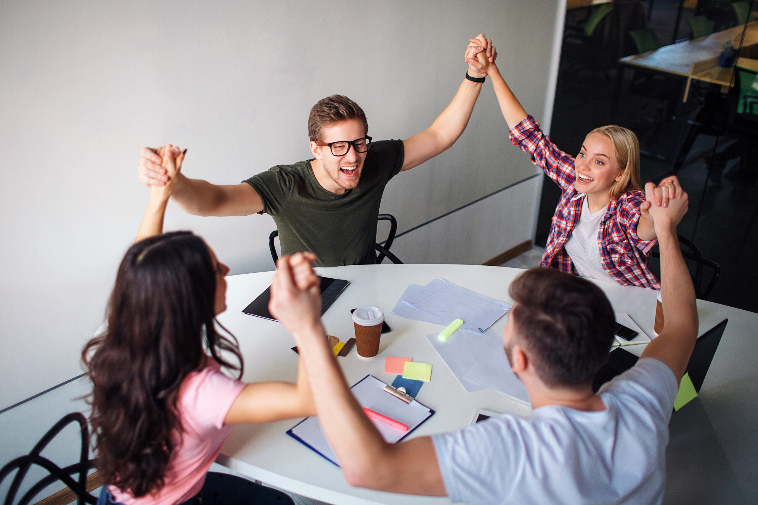 Happy young man and woman hold each other hands up and joying. They sit around white table in room. Team rejoicing.